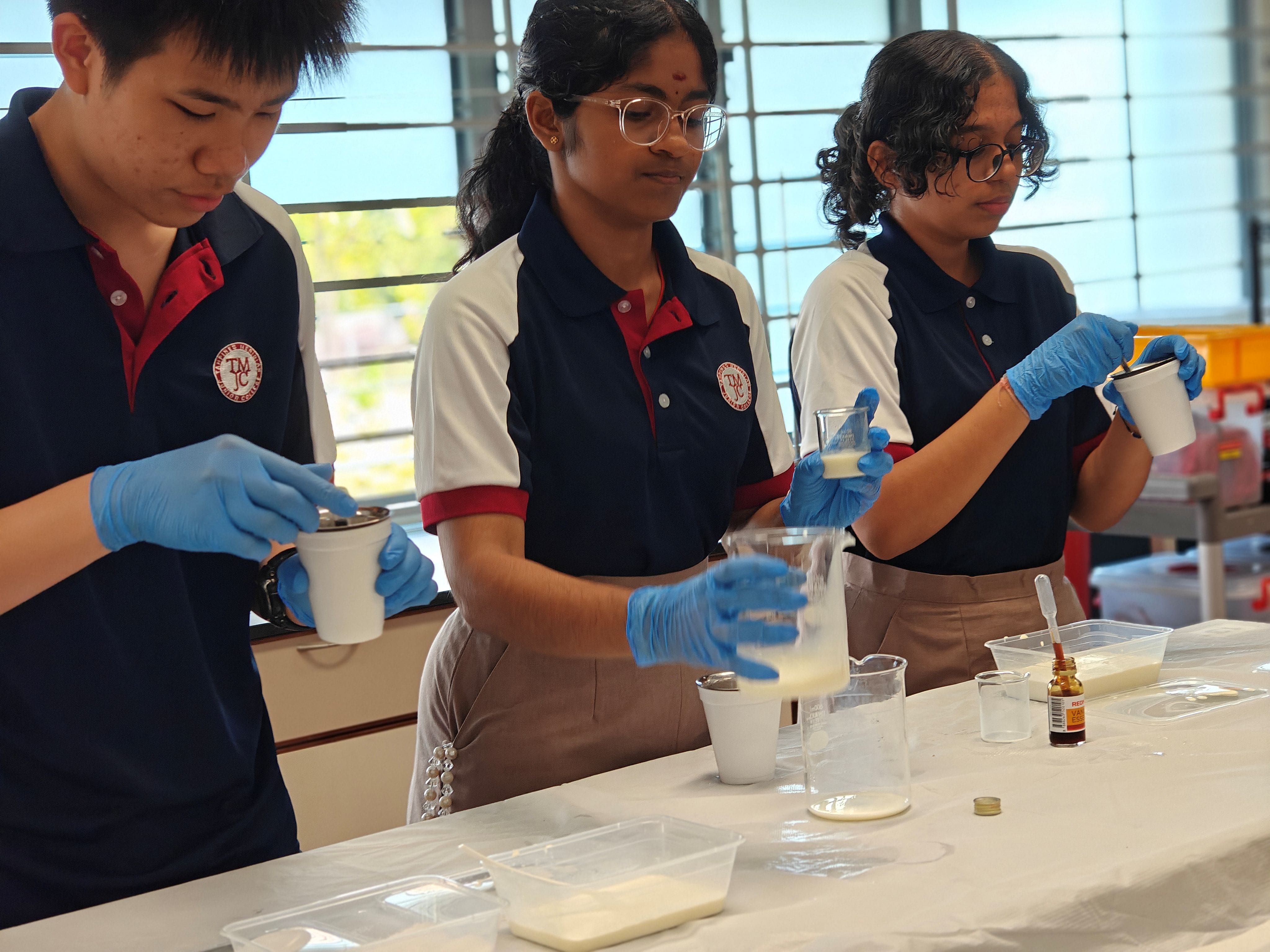 Three students in school uniforms and blue latex gloves carefully pour and mix liquids using beakers and cups on a lab bench, preparing ice cream ingredients.